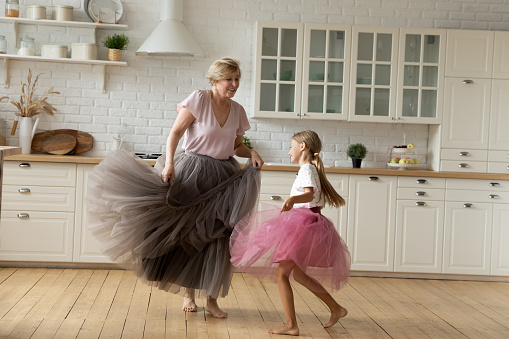 Merry leisure time. Happy energetic grandmother teach ball dance active little child. Caring grandma junior girl grandkid engaged in dancing in funny large fluffy skirts barefoot on warm kitchen floor