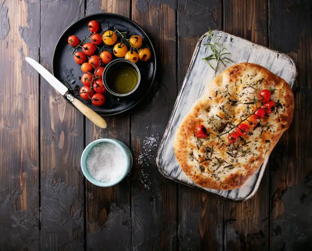 Home made rosemary focaccia flat bread served with baked tomatoes, olive oil and fresh rosemary, sea salt and knife over a wooden background. Top View