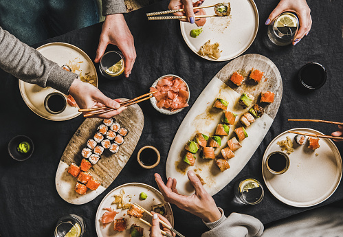 Family quarantine Japanese sushi dinner from delivery service, takeaway at home. Flat-lay of salmon, crab, prawn, vegan rolls, wasabi, ginger and people eating together over dark background, top view