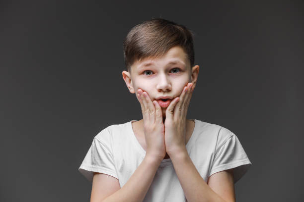 istock surprised boy holding his head on face isolated on gray background with copy space for text, child in shock, toothache. teenager in white t shirt 1305180854