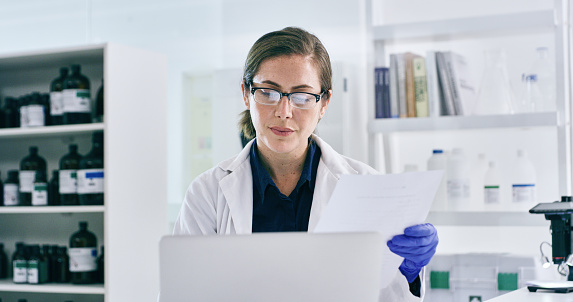 Shot of a young woman going through paperwork while working on a laptop in a lab