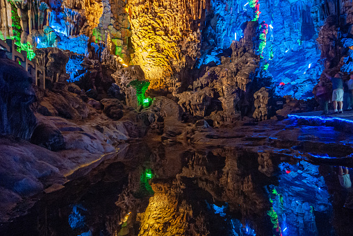 Guilin, China - May 11, 2010: Ludi Lu or Reed Flute Cave. blue lighted section of grotto with stalactites and stalagmites. Reflecting pond in front.