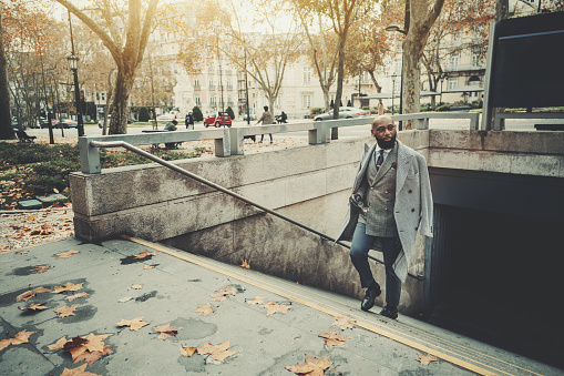 A dapper mature bald black man entrepreneur in a fashionable custom plaid costume and a coat, with a well-groomed beard, climbing the stairs on the street leaving the subway station and looking aside