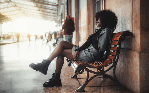 A side view of a tranquil young black African woman with curly African hair sitting relaxed on a wooden bench indoors of a covered station depot platform and waiting for the train