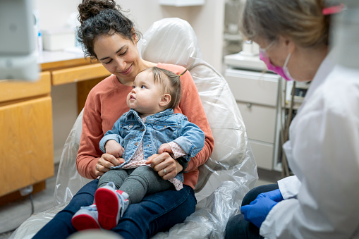 A young mixed race mother holds her calm one year old toddler in her lap during a consultation with the child's pediatric dentist.