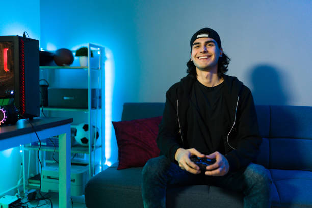 Excited man in his 20s having fun with a remote controller and a console Happy male gamer smiling while playing and winning in a video game with a remote controller. Young man sitting on the couch in his bedroom and enjoying a video game neon bedroom stock pictures, royalty-free photos & images