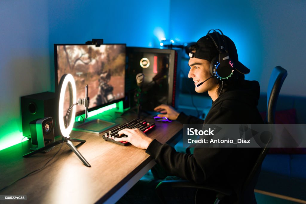 Latin man gaming on his PC during a live stream Smiling young man live streaming his online video game using a smartphone and a ring light. Happy gamer ready to start playing in a gaming computer Video Game Stock Photo