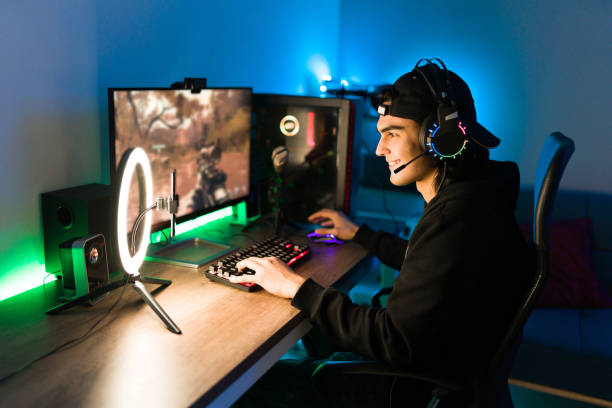 Latin man gaming on his PC during a live stream Smiling young man live streaming his online video game using a smartphone and a ring light. Happy gamer ready to start playing in a gaming computer games console stock pictures, royalty-free photos & images