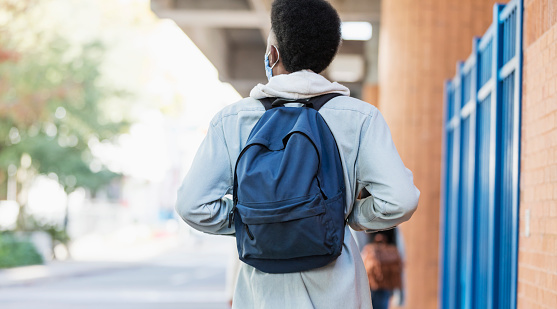 Rear view of a young African-American man in his 20s walking on a city sidewalk, carrying a backpack, wearing casual clothing. He is wearing a protective face mask, trying to prevent the spread of coronavirus during the COVID-19 pandemic.
