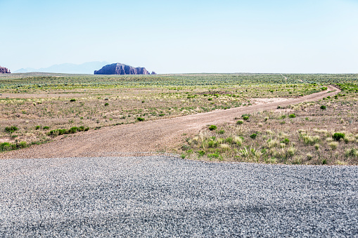At the end of a remote, deserted asphalt gravel highway, an empty winding dirt road stretches off into the distant prairie wilderness of the San Rafael Desert toward a distant rocky outcrop butte in the western USA state of Utah.