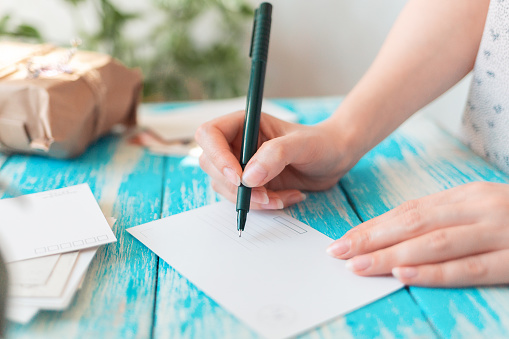 A woman writes on an empty postcard. Hands holding a pen close-up. Blue wooden table with a parcel in the background. The concept of mail correspondence and postcrossing.