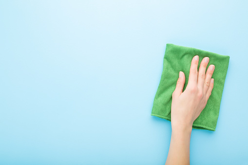 Young adult woman hand holding green rag and wiping table, wall or floor surface in kitchen, bathroom or other room. Empty place for text or logo. Light blue background. Pastel color. Top down view.