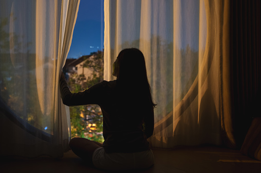 Rear view image of a woman opening bedroom curtain and looking outside the window at night