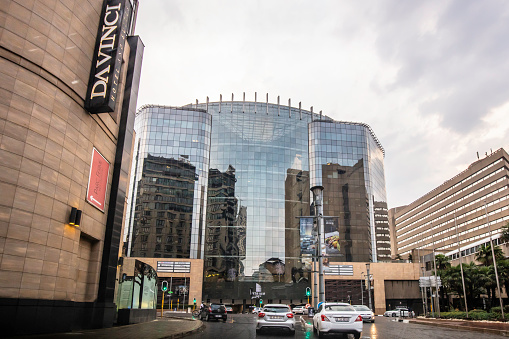 Sandton city centre with da vinci and intercontinental hotels, pedestrians, traffic at an intersection, under a cloudy raining season.