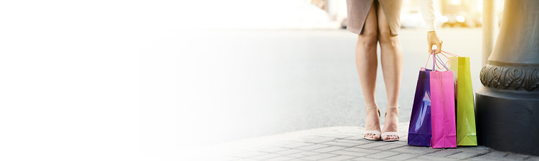 Woman shopping concept. Color paper bags in female hand on street, woman legs on heel shoes. Long banner