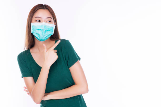 Positive young woman wearing safety medical mask protection from inhaling airborne bacteria or virus disease and air pollution problem on nose while pointing finger over isolated white background. mujer joven positiva con protección de máscara médica de seguridad contra la inhalación de bacterias en el aire o enfermedad del virus y problema de contaminación del aire en la nariz mientras señala con el dedo sobre fondo blanco aislado.