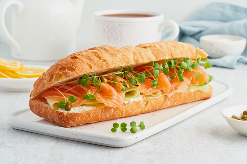 Big sandwich with salmon, cream cheese, cucumber slices on white concrete table. Morning healthy breakfast with fish and cup of drink, side view