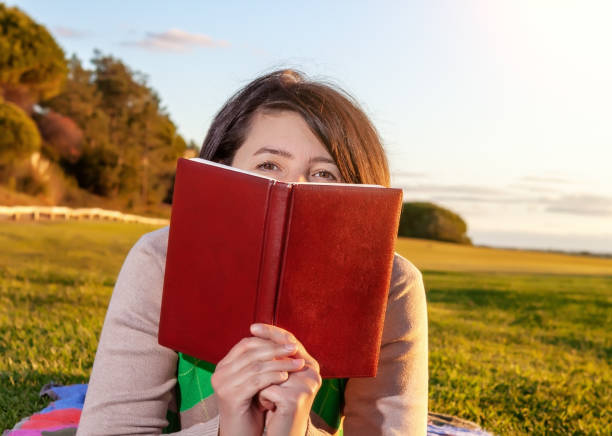 istock Middle aged woman peeking over book that she is holding, while lying down on some grass. Creamy sunset occurring in the background. 1305270963