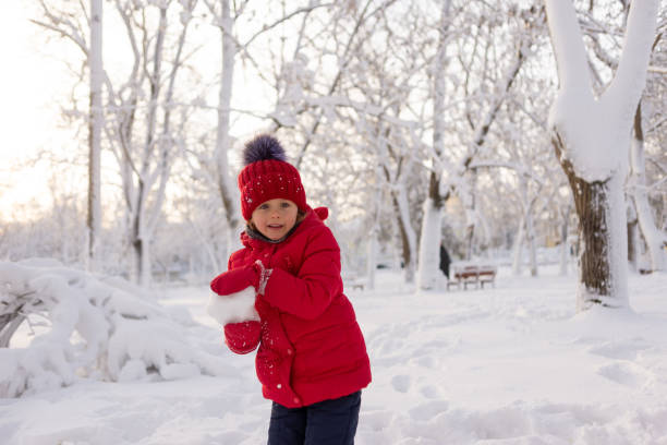 una niña de 6 años sola en manoplas, una chaqueta roja y un sombrero de punto en invierno se para en el parque y lanza bolas de nieve. juegos de invierno. - 6 7 years lifestyles nature horizontal fotografías e imágenes de stock