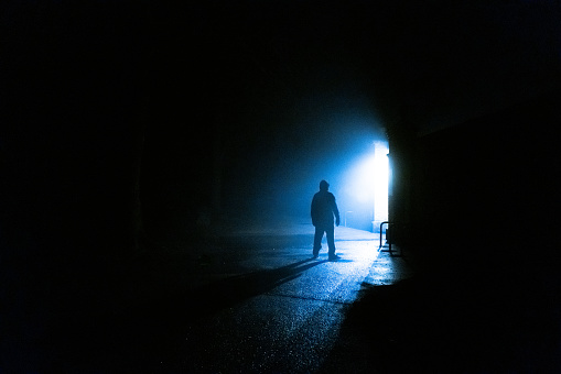 Moody dark image depicting a strange sinister man back lit by street lamps in the forest, rendering him a silhouette.