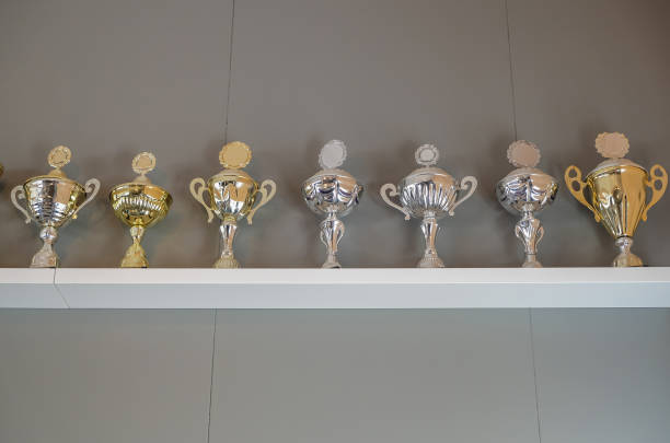 Shot of a group of silver and gold trophies from sports competitions arranged on top of a white shelf.