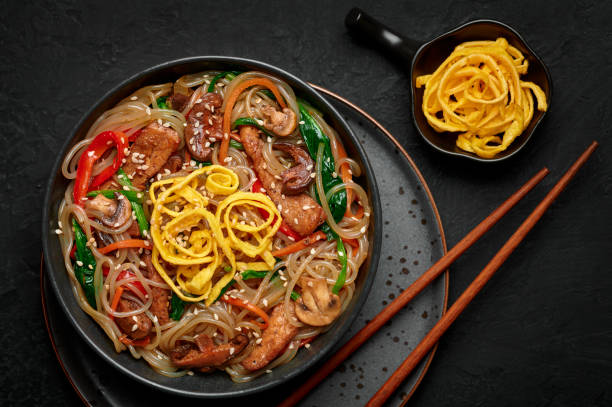 istock Japchae in black bowl on dark slate table top. Korean cuisine glass chapchae noodles dish with vegetables and meat. Asian traditional food. Authentic meal. Top view 1305283473