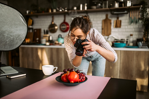 Young female photographer standing over food set, holding camera, photographing different types of fruit
