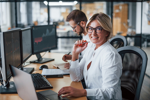 Woman looks into the camera. Two stockbrokers in formal clothes works in the office with financial market.