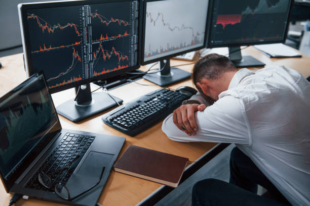 istock Tired businessman sleeping on the workplace by leaning on the table with multiple screens on it. Stock information on displays 1305289614