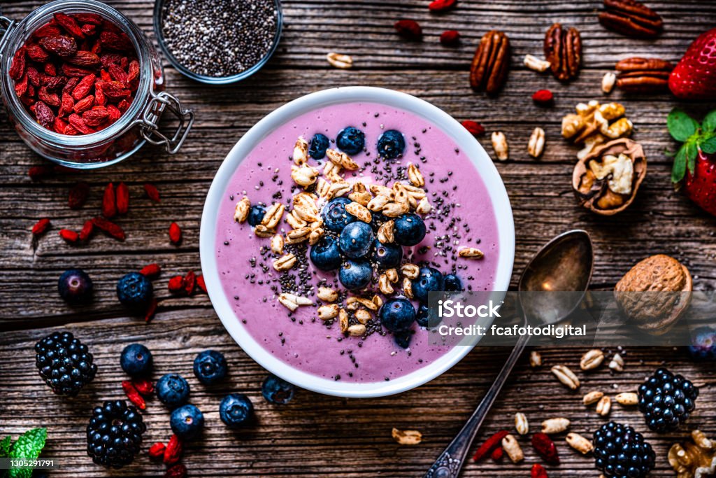 Mixed berries smoothie bowl on rustic wooden table. Healthy eating: overhead view of a mixed berries smoothie bowl with oat, chia seeds and nuts shot on rustic wooden table. Blackberries, blueberries, goji berries, pecan, walnuts and chia seeds are around the smoothie bowl. High resolution 42Mp studio digital capture taken with Sony A7rII and Sony FE 90mm f2.8 macro G OSS lens Smoothie Stock Photo