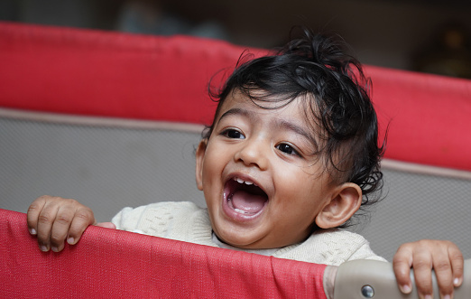 A closeup photo of an adorable indian toddler baby boy smiling with dimple in cheeks and standing inside a playpen.
