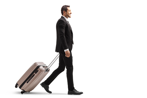 Full length profile shot of a young businessman in a suit walking and carrying a suitcase isolated on white background