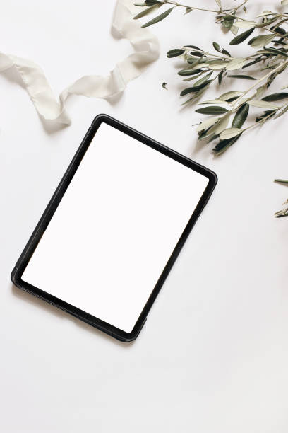 istock Electronic device mock-up. Closeup of tablet with empty screen. Olive tree branches and silk ribbon isolated on white table background. Flat lay, top view. Wedding design. Home office. Vertical. 1305302918