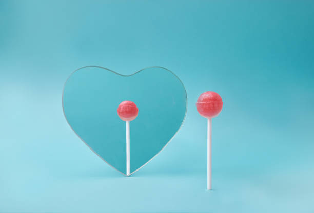 Self reflection concept with pink lollipop looking in heart mirror on blue background. Minimal front view.