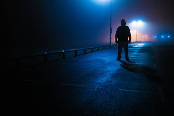 Sinister silhouette man lurking in deserted parking lot Moody dark image depicting a strange sinister man back lit by street lamps in an abandoned parking lot. creepy stalker stock pictures, royalty-free photos & images