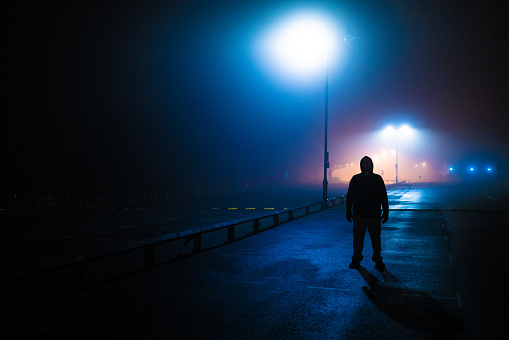 Moody dark image depicting a strange sinister man back lit by street lamps in an abandoned parking lot.