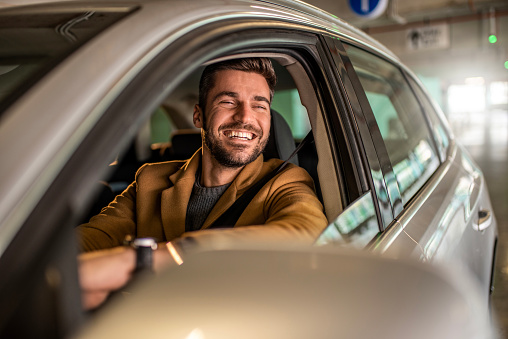 Photo of cheerful mid adult man driving a car in public garage.