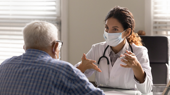 Close up female therapist wearing face mask consulting mature patient at meeting in hospital office, doctor talking, explaining, discussing medical checkup result or symptoms with older man