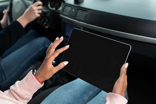 Urgent work, looking for address, gadget and traveling together. Young hands of african american female use tablet with empty screen, in passenger seat in car with driver, copy space, pov, cropped