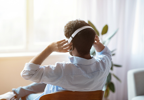 Unrecognizable Man Listening Music In Wireless Headphones At Home And Looking To Window, Brunette Guy Relaxing In Comfortable Chair And Enjoying His Favorite Melodies, Rear View With Copy Space