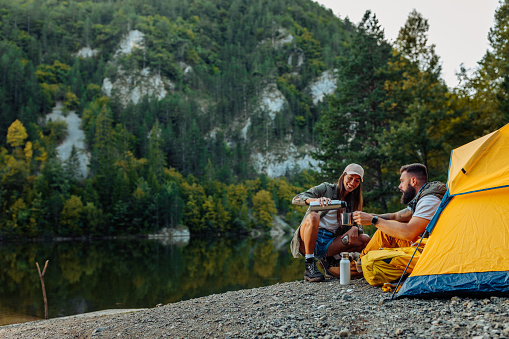 Couple sitting in front of their tent next to lake and drinking coffee