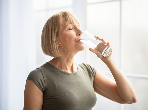 Fit senior woman drinking clear water during her workout break at home. Mature Caucasian lady staying hydrated after sports training. Healthy lifestyle and wellness concept