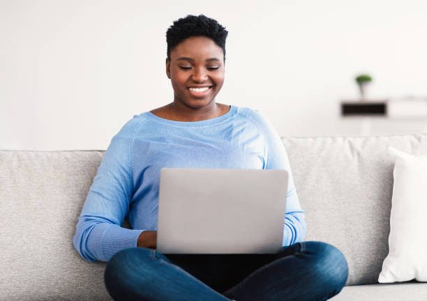 istock Smiling black woman using laptop, sitting on couch 1305329579