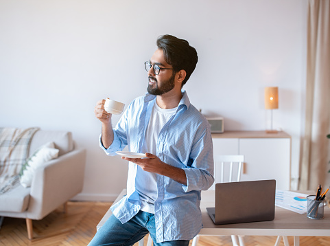 Handsome Eastern Man Relaxing With Cup Of Coffee After Working At Home Office, Smiling Millennial Arab Freelancer Guy Leaning Desk With Laptop, Enjoying Remote Work Opportunities, Copy Space