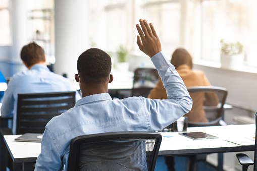 Education Concept. Rear back view portrait of african american guy student raising hand for answer or asking question, sitting at desk wearing surgical face mask at lecture in university or college