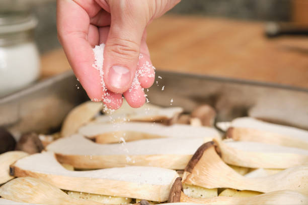 スライスされたキングトランペットキノコ(pleurotus eryngii)、ジャガイモ、タマネギの上に塩を加える人間の手のクローズアップ。調理プロセス。地中海の食事。 - 塩をふる ストックフォトと画像