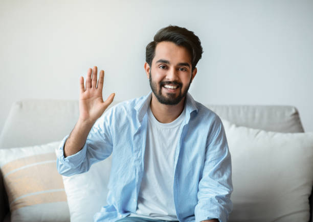 istock Greeting. Friendly eastern man waving hello while sitting on couch at home 1305330946