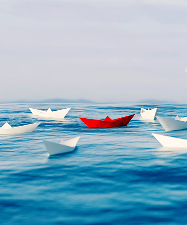 Concept image of standing out from the crowd. A red paper boat sails on the sea surrounded by white paper boats.
