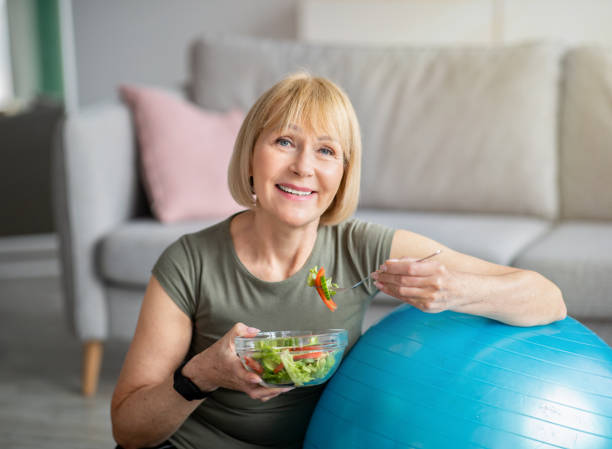 運動と健康的な食事の概念.自宅で新鮮な野菜サラダを食べるフィットボールを持つシニア女性 - sport food exercising eating ストックフォトと画像