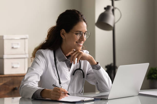 Smiling professional female doctor taking notes, looking at laptop screen Smiling professional female doctor wearing glasses and uniform taking notes in medical journal, filling documents, patient illness history, looking at laptop screen, student watching webinar telemedicine stock pictures, royalty-free photos & images